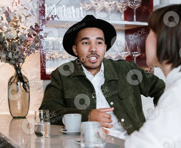 A man wearing a black hat and green jacket talking in a social setting.