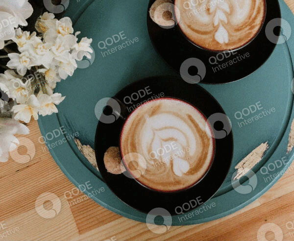 Latte art coffee in a black cup on a blue tray with cookies and flowers.