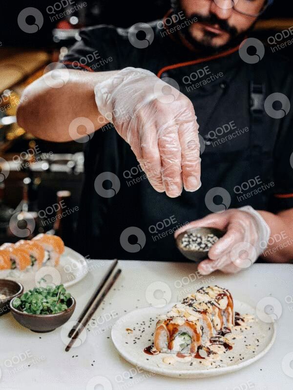Chef sprinkling seasoning on sushi rolls in a kitchen.