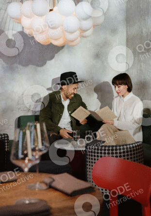 A man and woman in vintage attire share a book in a cozy, retro-styled room.