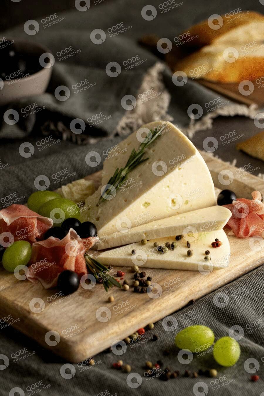 A cheese wedge garnished with herbs and surrounded by olives and prosciutto.