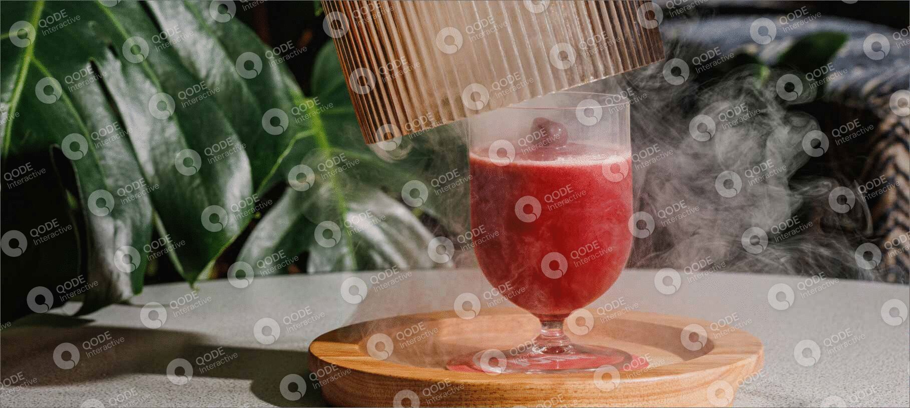 A glass of red smoothie on a wooden coaster with a green plant in the background.