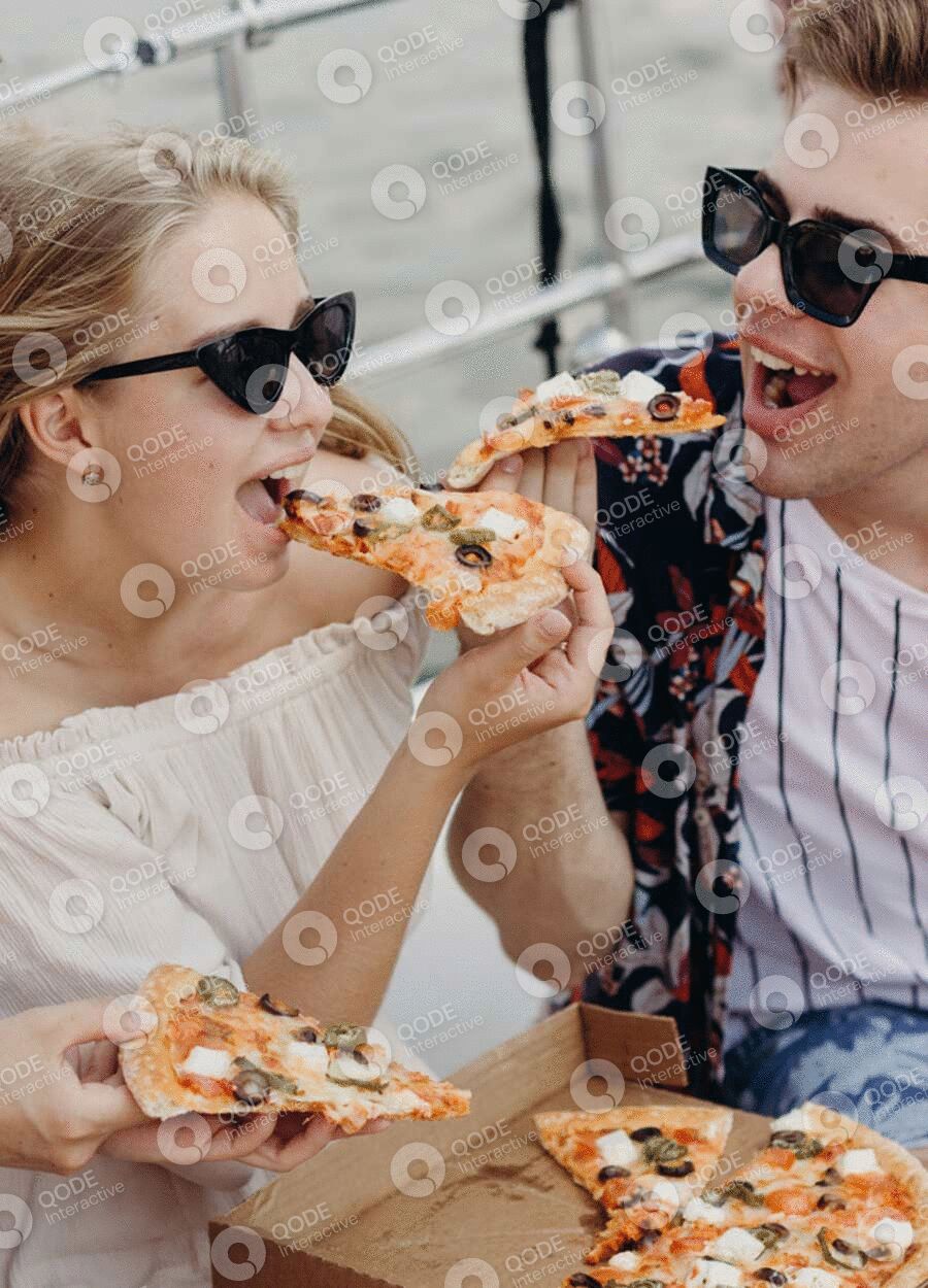 Couple joyfully sharing a slice of pizza outdoors wearing sunglasses.