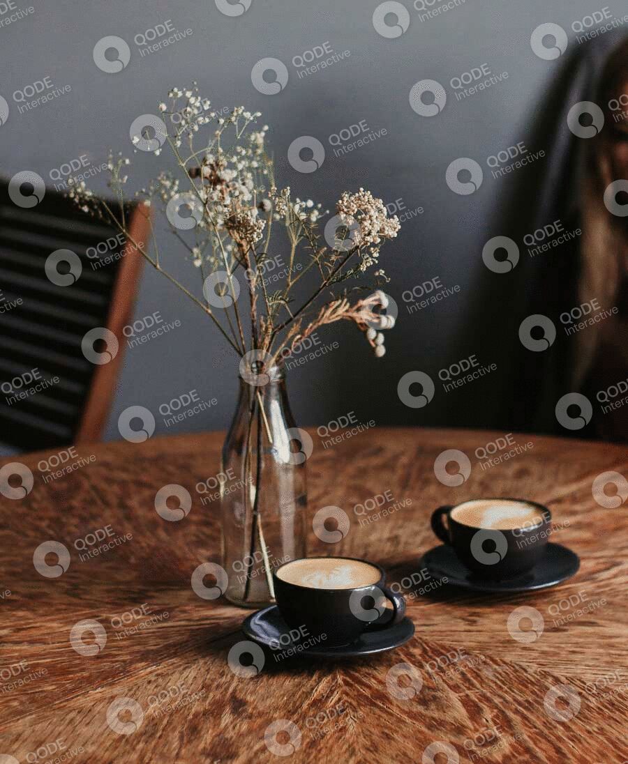 Two cups of coffee beside a vase of dried flowers on a wooden table.