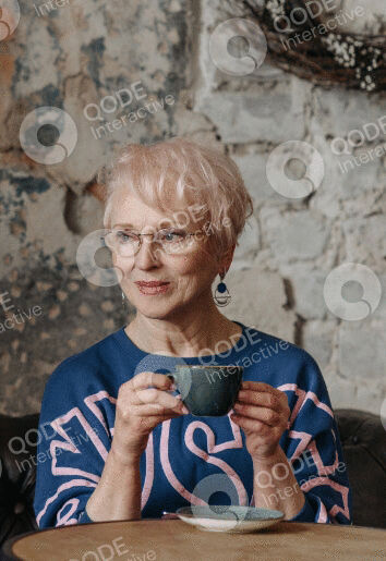 Smiling elderly woman holding a cup, wearing glasses and a blue sweater.