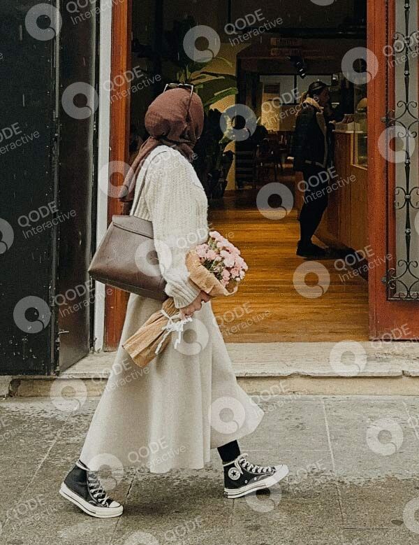 A person in a light coat holding flowers and a bag, walking past a shop.